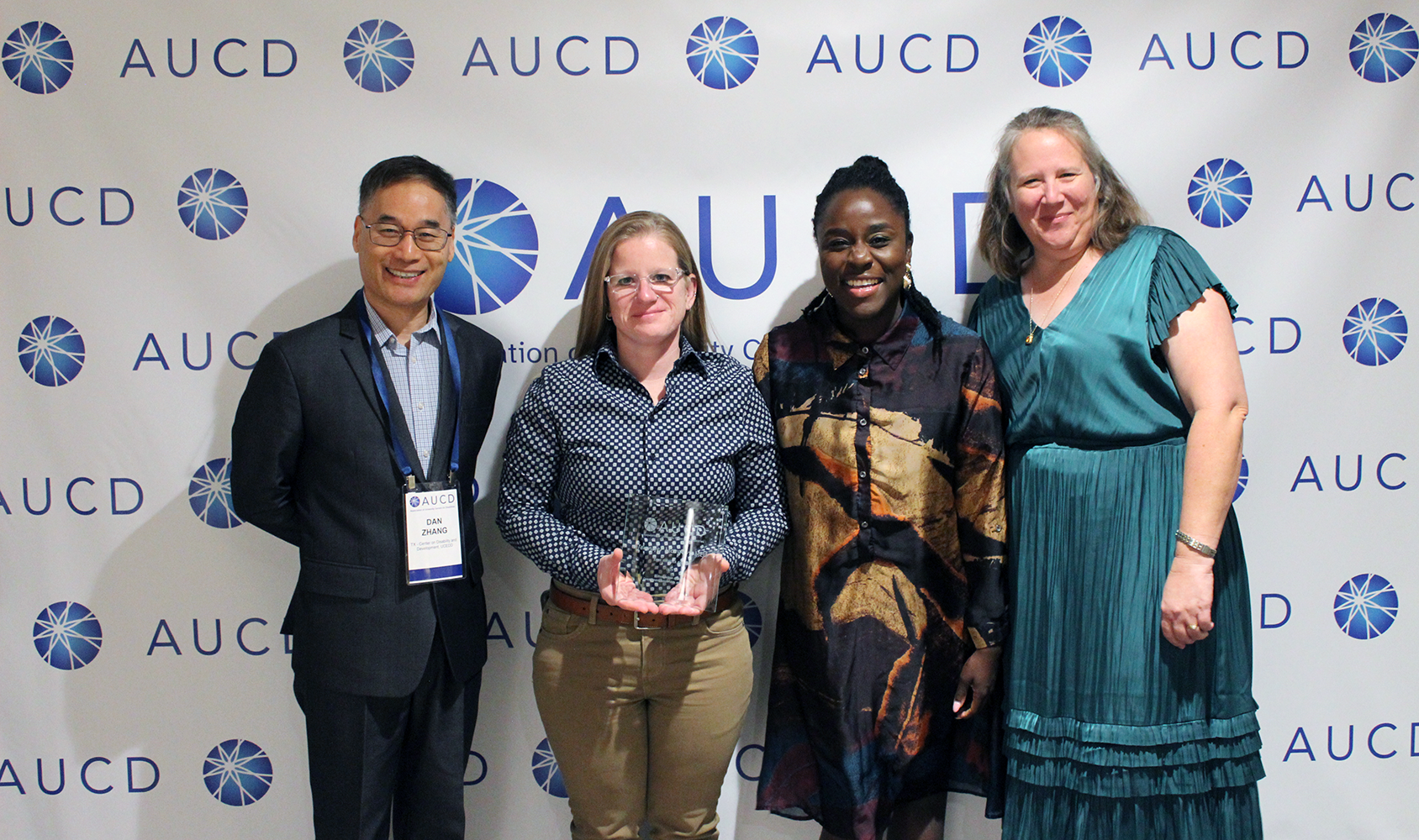 A conference panel scene shows two panelists seated behind a table with microphones and water bottles, with one panelist speaking into a microphone. Below the photo, a graphic reads “AUCD 2026 Annual Conference” with the theme “Impact through Innovation and Partnership,” and the dates “November 8–12 • Washington, DC,” alongside a circular blue conference logo.